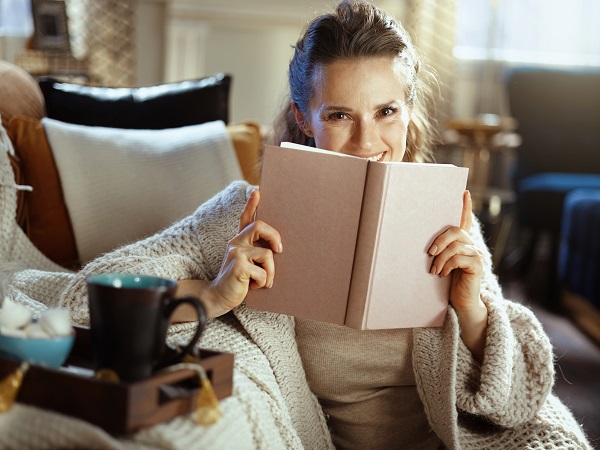 The image shows a woman relaxing at home, sitting comfortably on a couch wrapped in a cozy knit blanket. She is holding an open book close to her face, peeking over the top with a warm smile, suggesting that she is enjoying her reading. The atmosphere is cozy and inviting, with soft lighting that enhances the serene and peaceful ambiance of the room. A tray with a mug of tea or coffee and some marshmallows sits nearby, adding to the comfort of the setting. The overall vibe is one of warmth, relaxation, and contentment. Join Chapters club