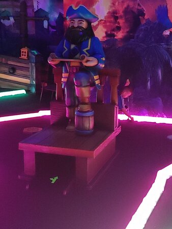 A pirate-themed glow-in-the-dark mini golf course featuring a statue of a pirate captain holding a scroll. The pirate is wearing a blue hat and jacket, standing on a wooden platform surrounded by neon-lit pathways. The course is illuminated with vibrant neon colors, enhancing the dark atmosphere of the room